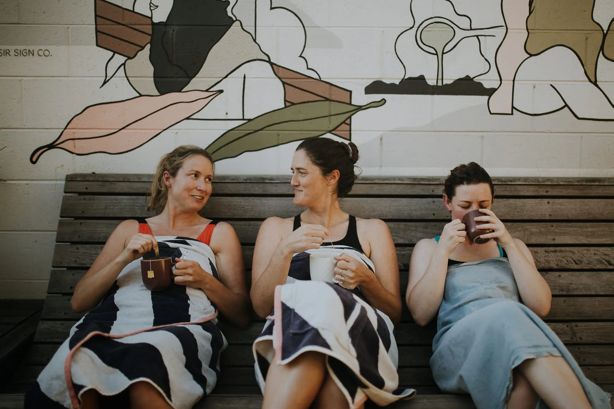 Three friends share tea on the bench beneath the bathers mural after a sauna