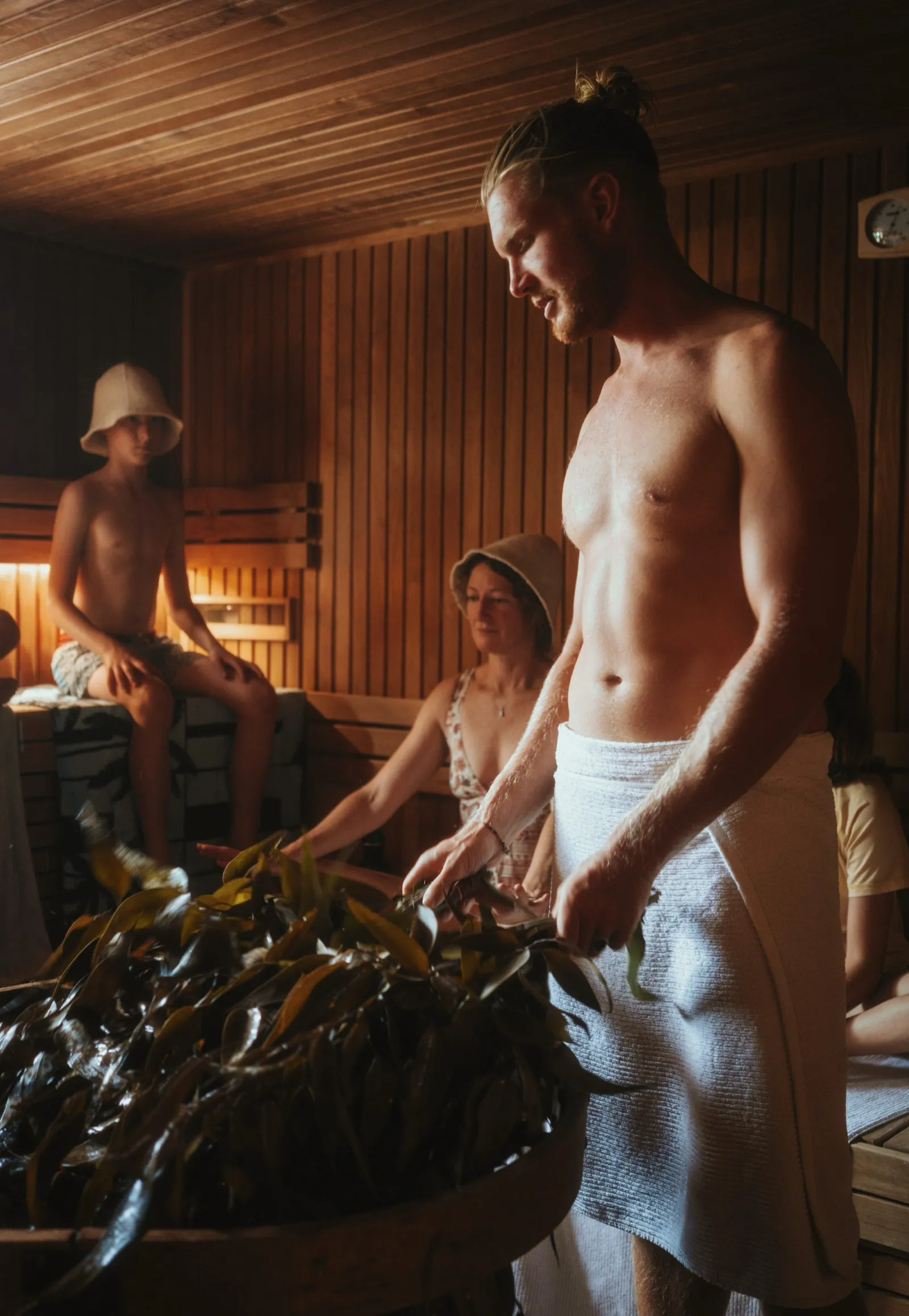 A Saunameister beats a eucalyptus whisk against the heater stones, water spraying off it, during an Aufguss ceremony with bathers seated around.