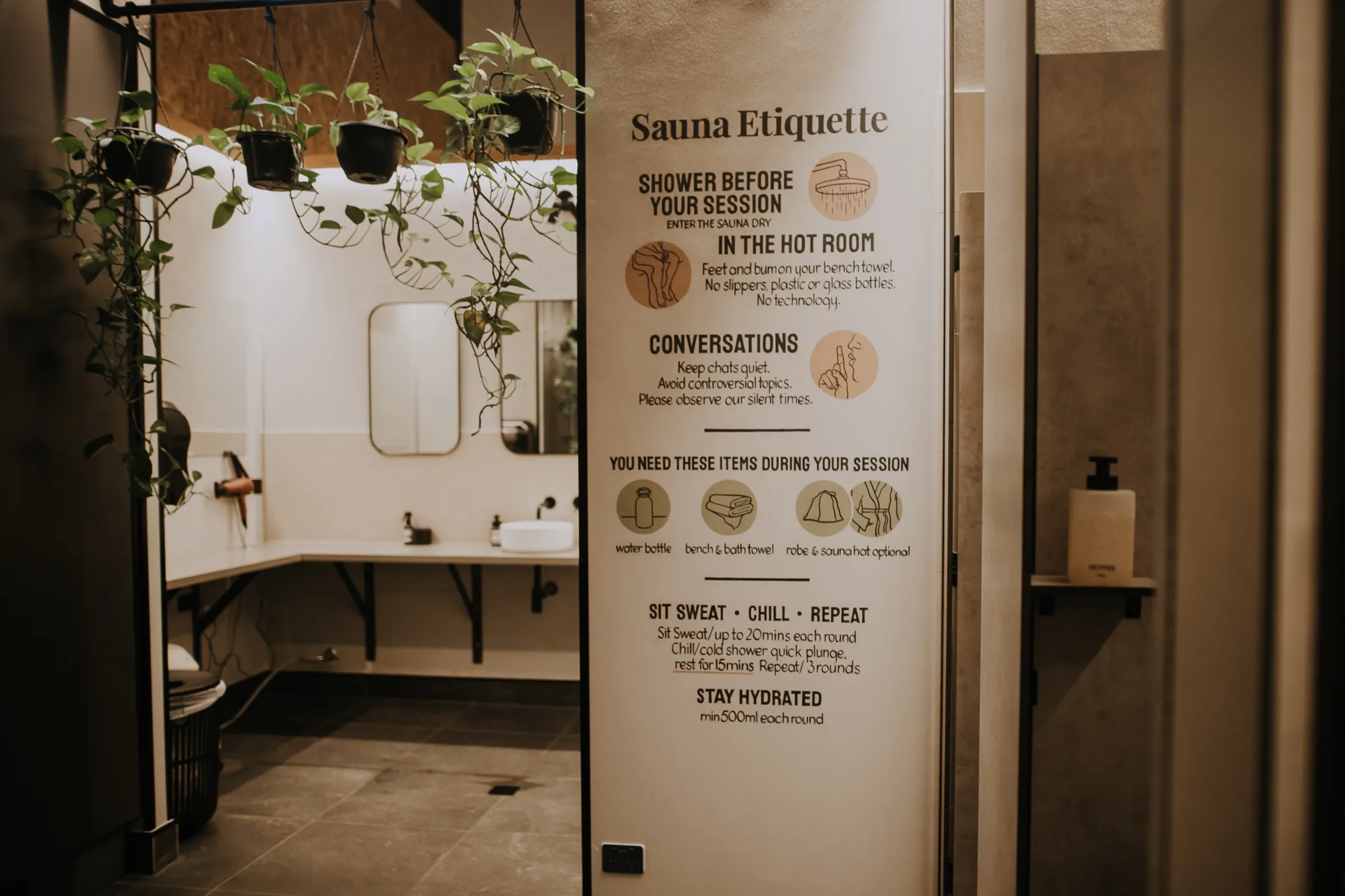 Changeroom vanity and mirror beside the Sauna Etiquette wall, framed by hanging plants