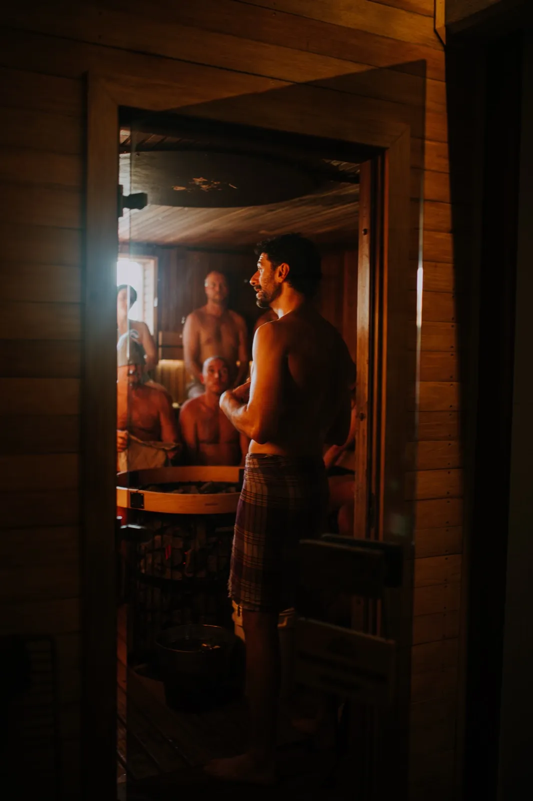 A Saunameister stands before the stove inside the sauna, bathers seated on the benches behind.