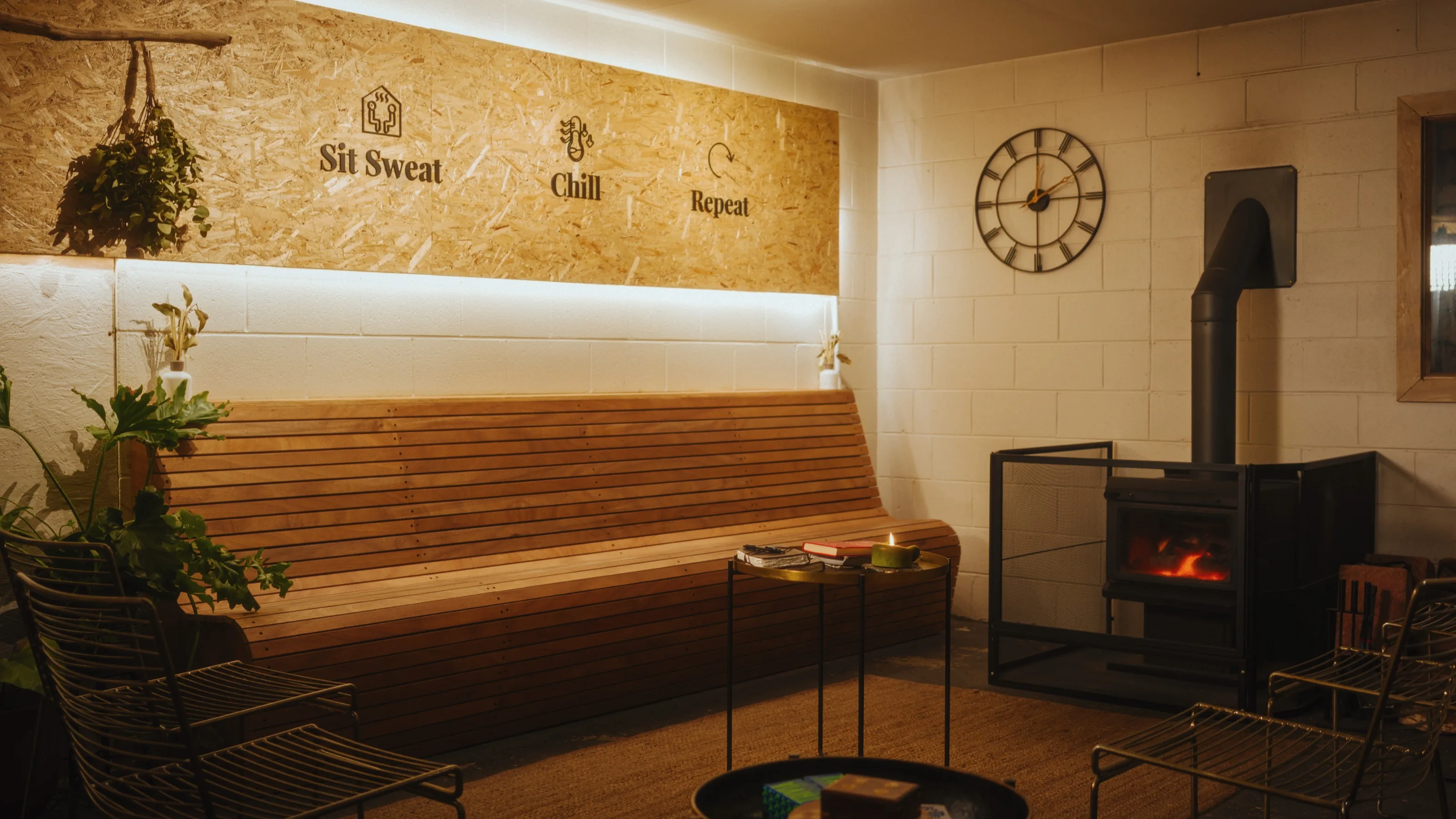 The upstairs rest lounge at Blue Mountains Sauna, with the Sit, Sweat, Chill, Repeat ethos on the timber wall and a wood fire glowing alongside the long bench.