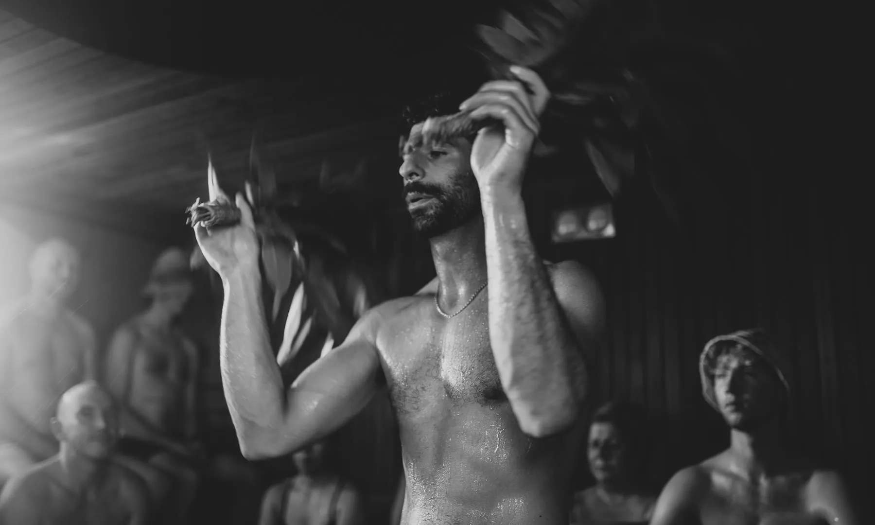 A Saunameister raises eucalyptus bundles overhead during an Aufguss, bathers around (black and white)